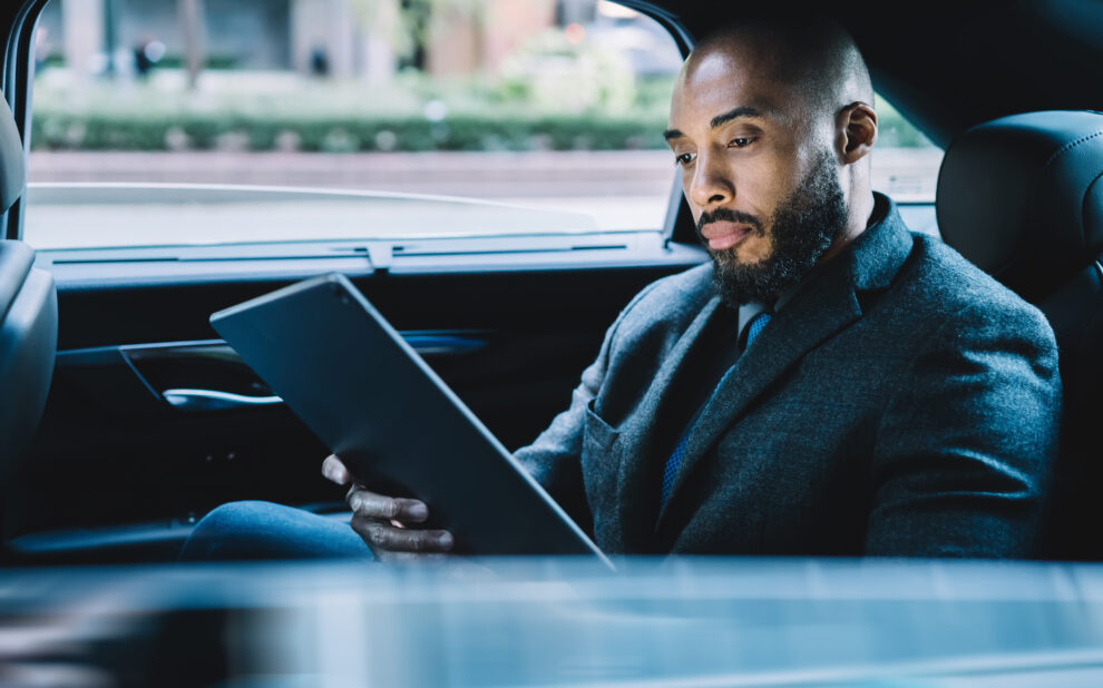 Businessman with black skin sitting on backseat in taxi automobile and searching contact number for making online video conference via modern touch pad, concept of market digitalisation on tablet