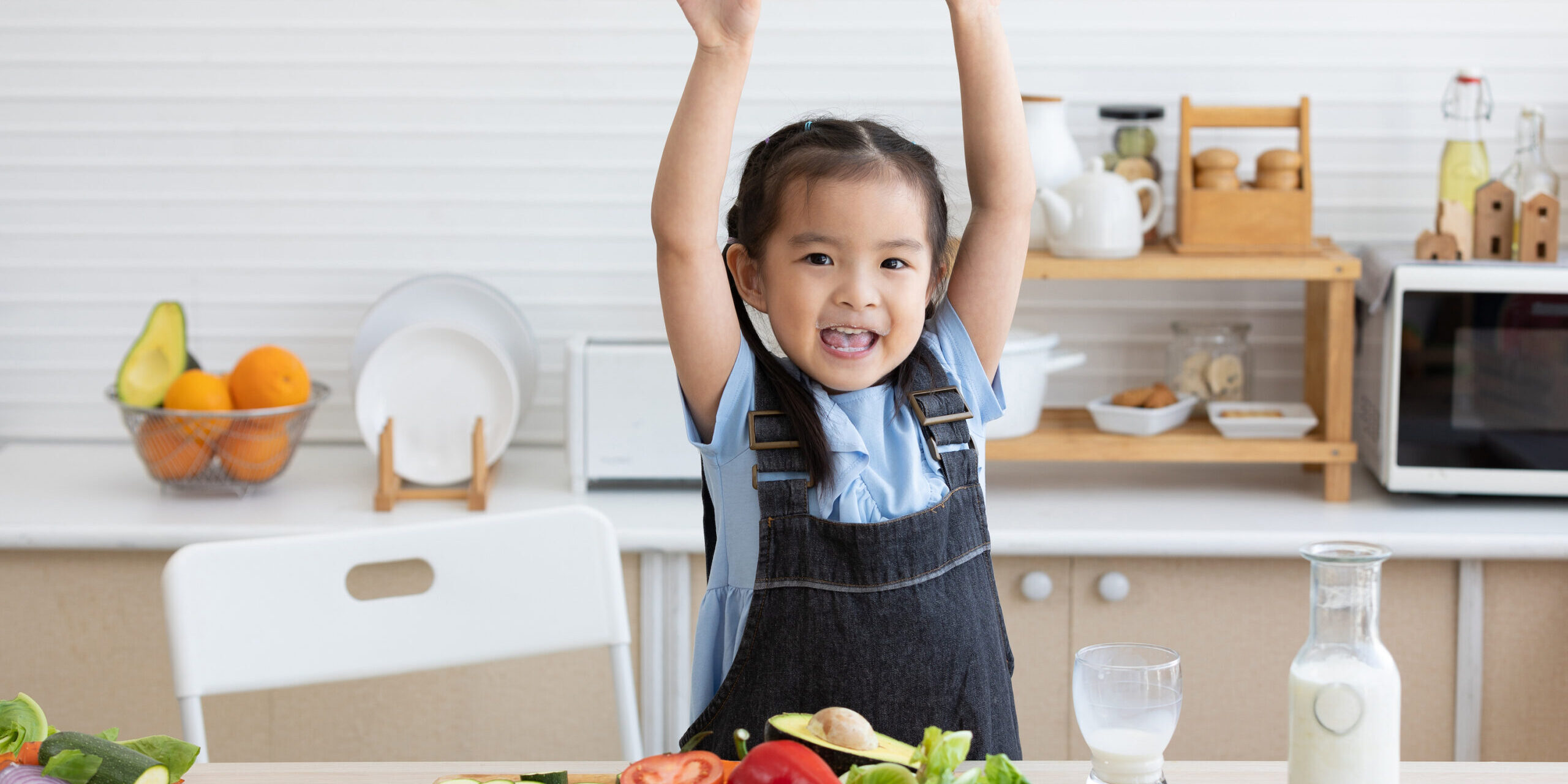 cute asian little child celebrating her success with raised arms pose after drinking milk and eating vegetables in the kitchen