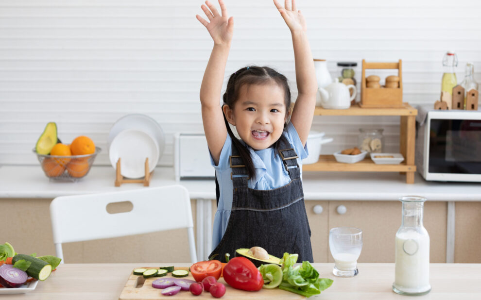 cute asian little child celebrating her success with raised arms pose after drinking milk and eating vegetables in the kitchen